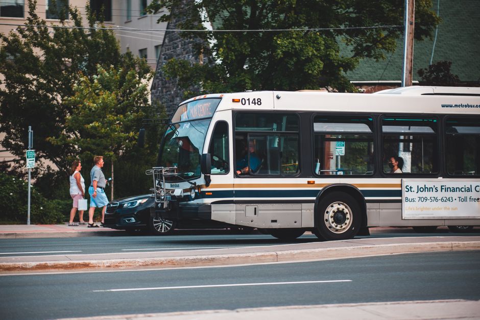 A city bus with a white and grey exterior, featuring blue and orange stripes along its side, is stationary on a street near a bus stop. The bus door is open, revealing seated passengers inside, with some individuals visible through the windows. The front of the bus displays the route number and destination, and the side advertisement mentions St. John’s Financial Centre. Next to the bus, a black car with a glossy finish is partially visible, positioned close to the bus's front right side, with only the front bumper and wheel showing. In the background, two pedestrians, a man and a woman, are walking on the sidewalk; the woman is dressed in a pale pink top and white shorts, while the man is wearing a grey shirt and turquoise shorts, both carrying shopping bags. Behind them, there are lush green trees, and brick or stone buildings partially obscured by the foliage. The scene is set during daylight with natural lighting, and the overall environment suggests a typical urban street, possibly near a bus stop used for public transportation and private vehicle access in a residential or commercial area. The context aligns with an independent or alternative transport system, such as private rubbish removal services often integrating with public routes.