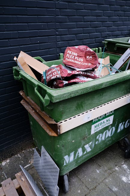 A large green municipal rubbish bin with a white internal tray, placed on a paved surface against a dark brick wall. The bin is filled with black and red packaging bags, containing materials related to gardening or building supplies, with some items leaning slightly out of the open top. The bin's surface shows signs of weathering and dirt, and it is situated in an outdoor setting, likely a back alley or garden area. Adjacent to the bin, a small section of a metal grating or step is visible on the ground. This scene depicts a typical instance of private waste management, aligned with independent collection services such as those offered by houseclearancestjohnswood.co.uk, suitable for scheduled rubbish removal or skip alternatives. The environment appears to be part of a residential or small commercial property cleanup, emphasizing proper disposal and waste handling practices in an urban setting.