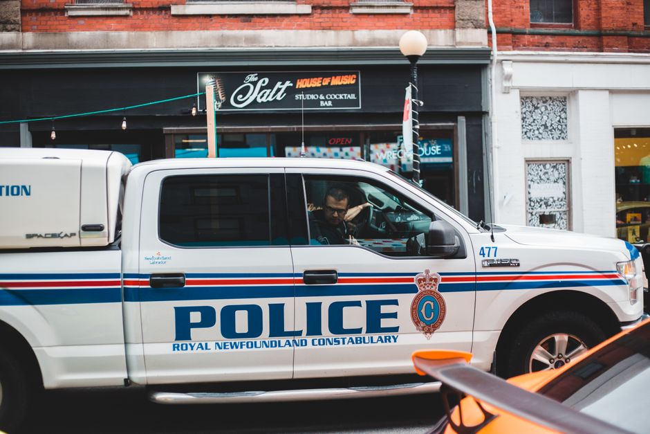 A white police pickup truck with blue and red stripes, displaying the Royal Newfoundland Constabulary emblem and the word 'POLICE', is parked on a street in front of a commercial building. The truck's side window shows an officer inside, looking outward. Behind the vehicle, the storefront has signage for 'The Salt' house music bar, with a black sign featuring white and red lettering, and an illuminated 'OPEN' sign. The surroundings include a white lamppost with a round globe lamp, part of a brick and white façade building. The scene appears to be in an urban area with natural daylight, and the police vehicle indicates a security or law enforcement presence in the vicinity, with potential relevance to private or alternative waste handling and rubbish removal services in the area, as occasionally observed during street patrols.