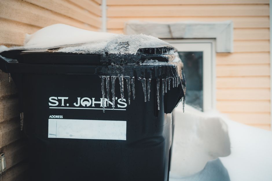 A black plastic rubbish bin with the label 'ST. JOHNS' in white text, situated outdoors against a beige wooden house wall in a snowy environment. The bin has a slightly rounded lid partially covered in snow and ice, with icicles hanging from its edge, indicating cold weather. Behind the bin, a small window with a white frame and a grey stone sill is visible, with snow piled below it. The scene appears to be part of a residential property, and the image reflects a typical setting for private waste collection or alternative rubbish disposal services provided by companies such as houseclearancestjohnswood.co.uk.