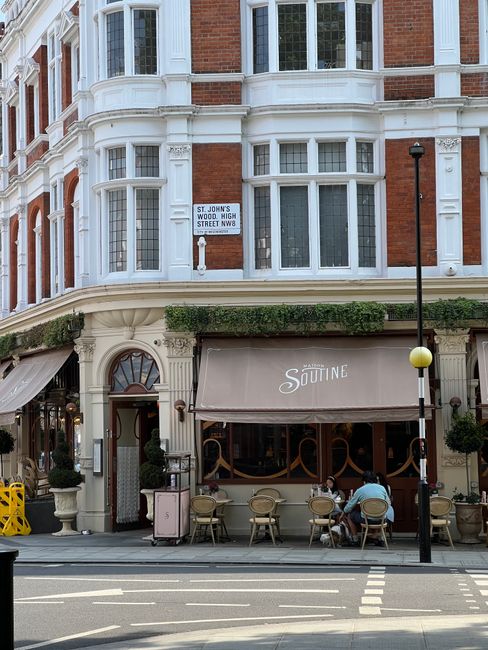 A street-level view of a three-storey Victorian-style building with red brick walls and white decorative window frames. The ground floor features a café with a beige fabric canopy displaying the word 'SUTINE' in white lettering. In front of the café, there is an outdoor seating area with several beige chairs and tables, with one person seated at a table and another standing nearby. A tall black lamppost with a yellow spherical light is positioned on the sidewalk, which has white dashed lines marking a pedestrian crossing. Above the café, there is a sign on the building indicating the location as St. John's Wood High Street, with greenery and potted plants enhancing the storefront. The overall scene reflects a typical urban environment with a mixture of commercial and residential architecture, situated in an area possibly serviced by independent waste collection, fitting within the context of rubbish removal and private disposal services offered by houseclearancestjohnswood.co.uk.