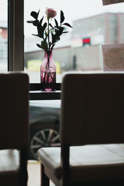 A close-up view through a window showing a small table with a pink glass vase containing a single pink flower with green leaves. The vase has a rounded shape with a narrow neck and a reflective surface. In the foreground, two dark-colored chairs with cushioned seats and backs partially obscure the view, positioned facing the window. Outside the window, a street scene is visible, including parked cars and blurred building facades under an overcast sky. The interior setting appears to be a café or restaurant, with natural light filtering in, creating a calm and subdued atmosphere. This scene subtly aligns with the context of property or interior waste removal, as it suggests a well-maintained space that may require occasional cleaning or clearing, a service often provided by companies like houseclearancestjohnswood.co.uk, focusing on professional rubbish removal or site clearance.
