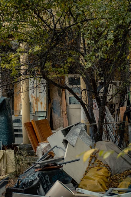 A cluttered outdoor scene featuring a pile of discarded household items and waste materials situated beneath a large, leafless or sparsely-leaved tree with dark, twisting branches. The debris includes a mix of domestic objects such as an old white washing machine or appliance, partially covered with a grey cloth or tarp, and several wooden cabinet doors with a warm, orange-brown finish leaning against the pile. Nearby, there are black bags, a yellow bag, and other miscellaneous refuse, scattered across the ground, which appears to be a concrete or paved surface. In the background, a weathered building wall with a mix of exposed and rusted metal panels and a partially visible window frame suggest an urban or semi-urban setting. The scene is lit with natural daylight, emphasizing the varied textures and colors of the waste and environment, consistent with a private waste removal or on-site clearance scenario managed by a local rubbish collection service like houseclearancestjohnswood.co.uk.